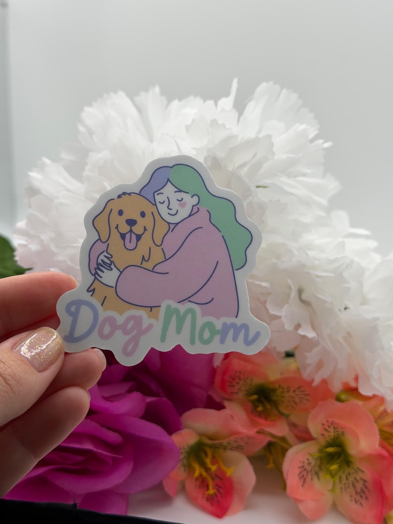 Op de afbeelding: Een gestanste sticker met een kleurrijke illustratie van een persoon met lichtblauw en groen haar in een roze trui, die een goudkleurige hond knuffelt. De tekst "Dog Mom" is onderaan de sticker gedrukt in een lichtpaars cursief lettertype.