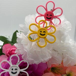 Peut inclure: Trois porte-cl&eacute;s en forme de fleur de couleur rose, jaune et blanche. Chaque porte-cl&eacute;s pr&eacute;sente un visage circulaire central avec un sourire clin d'&oelig;il noir et six p&eacute;tales arrondis. Un anneau en m&eacute;tal argent&eacute; est fix&eacute; au sommet de chaque fleur.