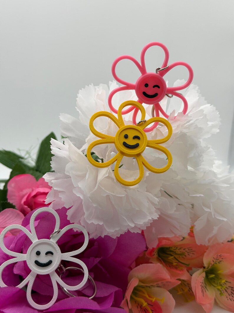 Peut inclure: Trois porte-cl&eacute;s en forme de fleur de couleur rose, jaune et blanche. Chaque porte-cl&eacute;s pr&eacute;sente un visage circulaire central avec un sourire clin d'&oelig;il noir et six p&eacute;tales arrondis. Un anneau en m&eacute;tal argent&eacute; est fix&eacute; au sommet de chaque fleur.