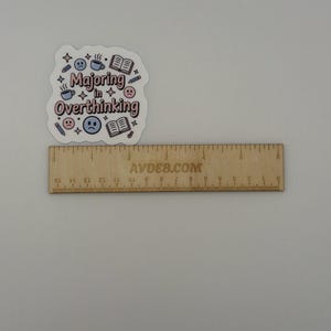 May include: A die-cut sticker with the text "Majoring in Overthinking" in pink and black, surrounded by blue and pink icons of books, coffee mugs, pencils, and sad faces, positioned above a wooden ruler measuring approximately 6 inches in length.
