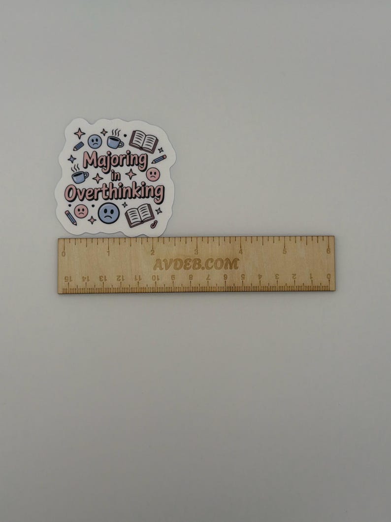 May include: A die-cut sticker with the text "Majoring in Overthinking" in pink and black, surrounded by blue and pink icons of books, coffee mugs, pencils, and sad faces, positioned above a wooden ruler measuring approximately 6 inches in length.