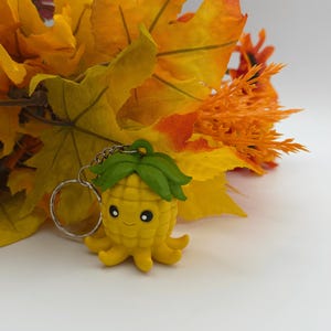 May include: A yellow pineapple-shaped octopus keychain with a green leafy top, black and white eyes, and a small smile. The figure has a textured, grid-like body and multiple tentacles at the base, attached to a silver metal split ring.