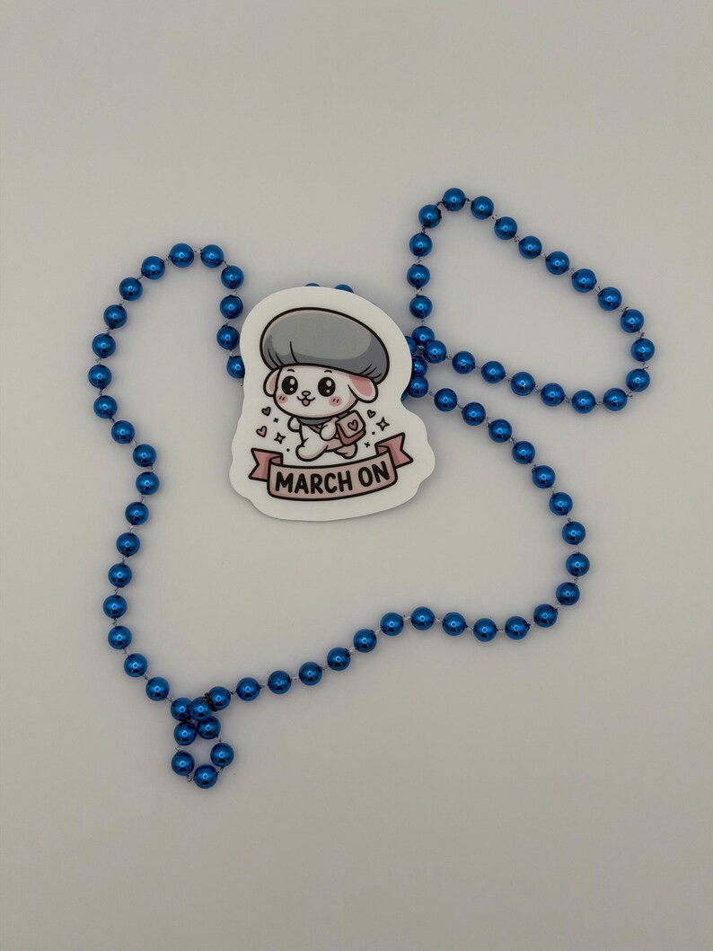 K&ouml;nnte beinhalten: Ein gestanzter Aufkleber mit einer Cartoon-Figur mit grauem pilzartigem Hut, einer rosa Umh&auml;ngetasche und einem rosa Banner mit dem Text MARCH ON, umgeben von einer langen Kette aus gl&auml;nzenden, metallisch blauen, kugelf&ouml;rmigen Perlen.