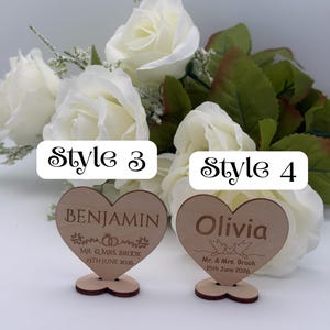 May include: Two heart-shaped wooden signs with stands. The left sign reads Style 3, BENJAMIN, MR. & MRS. BROOK, 15TH JUNE 2026 with ring graphics. The right sign reads Style 4, Olivia, Mr. & Mrs. Brook, 15th June 2026 with bird graphics.