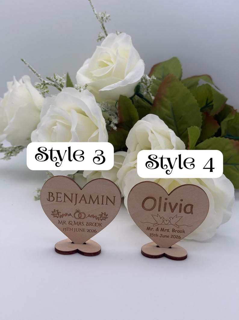 May include: Two heart-shaped wooden signs with stands. The left sign reads Style 3, BENJAMIN, MR. & MRS. BROOK, 15TH JUNE 2026 with ring graphics. The right sign reads Style 4, Olivia, Mr. & Mrs. Brook, 15th June 2026 with bird graphics.