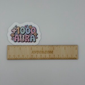 May include: A die-cut sticker featuring the text +1000 AURA in a bubble font with pastel colors, surrounded by small star and heart icons. The sticker is positioned above a wooden ruler that measures approximately 6 inches in length.