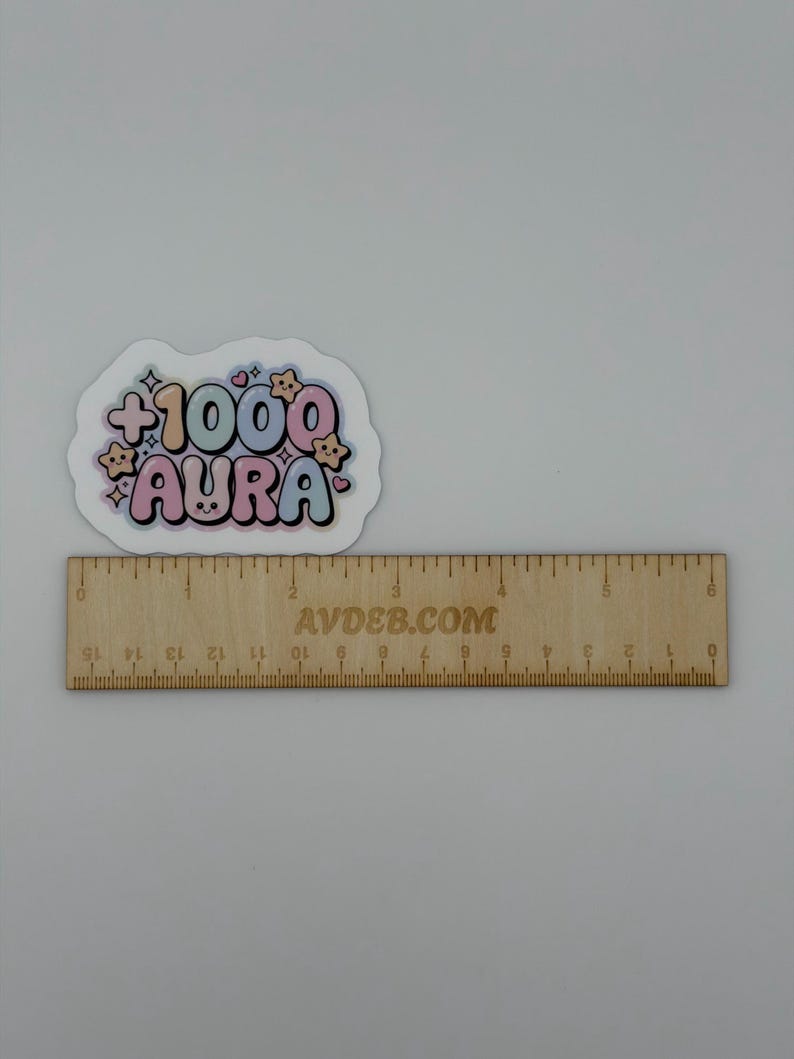 May include: A die-cut sticker featuring the text +1000 AURA in a bubble font with pastel colors, surrounded by small star and heart icons. The sticker is positioned above a wooden ruler that measures approximately 6 inches in length.