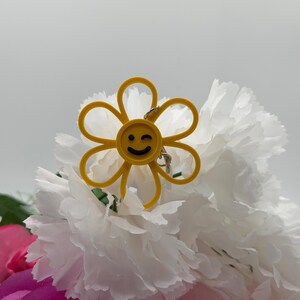 Peut inclure: Un porte-cl&eacute;s jaune en forme de fleur avec un visage circulaire central affichant un clin d'&oelig;il noir. Les p&eacute;tales sont d&eacute;limit&eacute;s par un fin contour jaune et un petit anneau m&eacute;tallique argent&eacute; est fix&eacute; au sommet de la fleur.