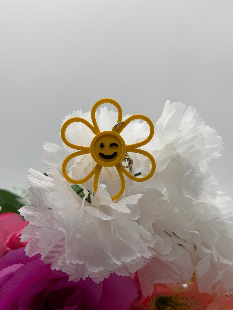 Peut inclure: Un porte-cl&eacute;s jaune en forme de fleur avec un visage circulaire central affichant un clin d'&oelig;il noir. Les p&eacute;tales sont d&eacute;limit&eacute;s par un fin contour jaune et un petit anneau m&eacute;tallique argent&eacute; est fix&eacute; au sommet de la fleur.