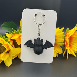 May include: A black bat keychain featuring a rounded body, pointed ears, and outstretched wings. The bat is attached to a silver-colored metal split ring by a short chain, and it is displayed on a plain white rectangular card for presentation.