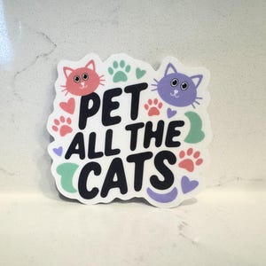 May include: A sticker with the text "PET ALL THE CATS" in black, with a pink cat and a purple cat at the top. The sticker also features paw prints, hearts, and a green swirl.