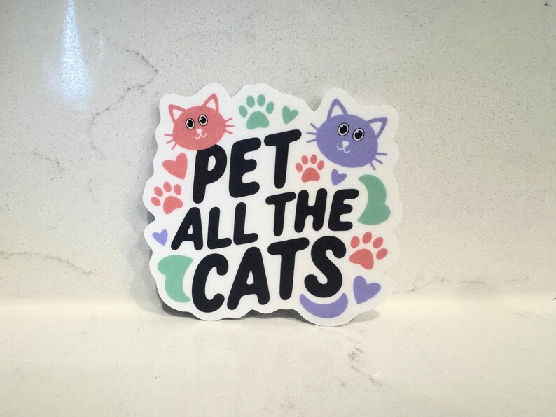 May include: A sticker with the text "PET ALL THE CATS" in black, with a pink cat and a purple cat at the top. The sticker also features paw prints, hearts, and a green swirl.