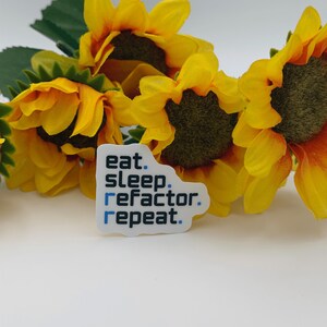 May include: A white rectangular sticker with rounded corners featuring the black text eat. sleep. refactor. repeat. with blue dots and a blue letter r. The sticker is positioned in front of three bright yellow artificial sunflowers with green leaves.