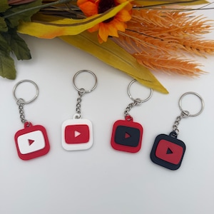 May include: Four square keychains featuring the YouTube play button logo. Two keychains have red bases with white centres, and two have black bases with red centres. Each keychain includes a silver metal split ring attached by a short chain.