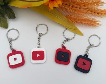 May include: Four square keychains featuring the YouTube play button logo. Two keychains have red bases with white centers, and two have black bases with red centers. Each keychain includes a silver metal split ring attached by a short chain.