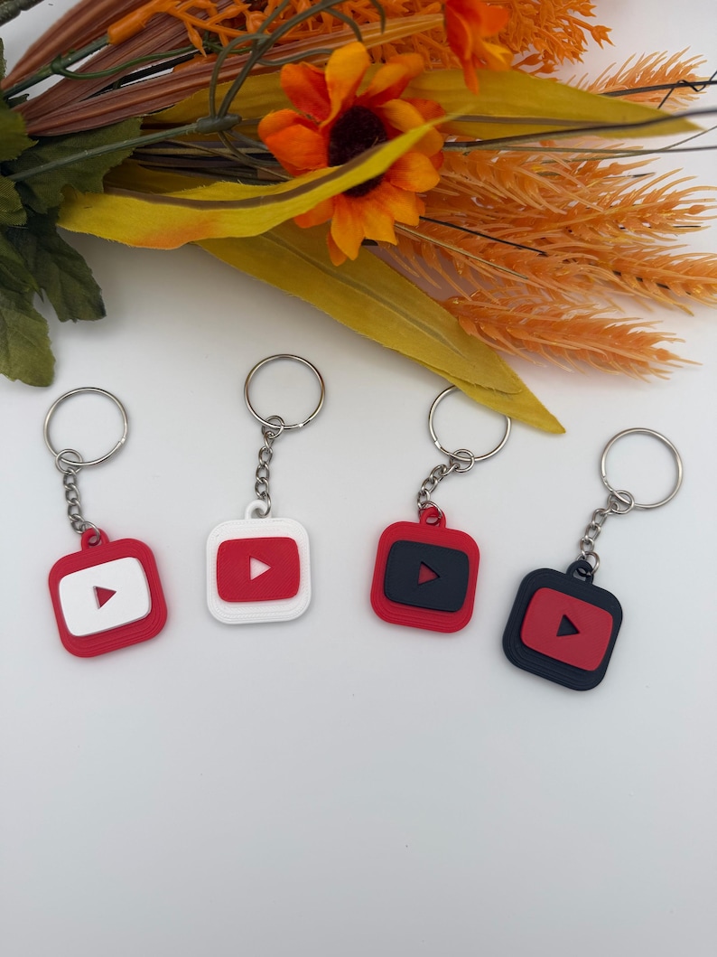May include: Four square keychains featuring the YouTube play button logo. Two keychains have red bases with white centers, and two have black bases with red centers. Each keychain includes a silver metal split ring attached by a short chain.