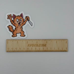 May include: A die-cut sticker featuring an orange tabby cat with an angry expression, holding a small silver knife in one paw. The sticker is positioned above a wooden ruler that measures 15 cm in length, with the text AVDEB.COM printed on it.