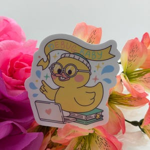 May include: A die-cut sticker featuring a yellow duck wearing round glasses and a beanie, sitting at a laptop with a heart icon. Above the duck is a yellow banner with the text DEBUG BABY. The design includes small blue and green accents.
