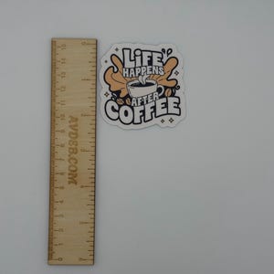 May include: A die-cut sticker featuring a coffee cup illustration with the text "LIFE HAPPENS AFTER COFFEE" in black and tan colours, positioned next to a wooden ruler showing a height of approximately 7.5 cm for the sticker design.