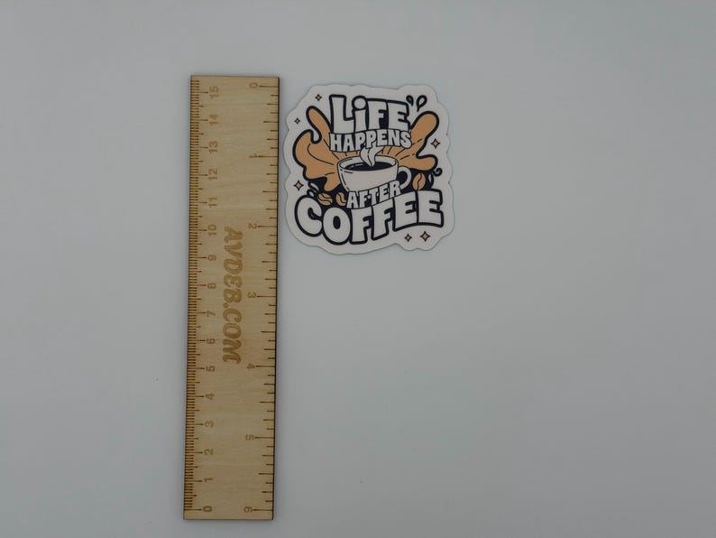 May include: A die-cut sticker featuring a coffee cup illustration with the text "LIFE HAPPENS AFTER COFFEE" in black and tan colours, positioned next to a wooden ruler showing a height of approximately 7.5 cm for the sticker design.