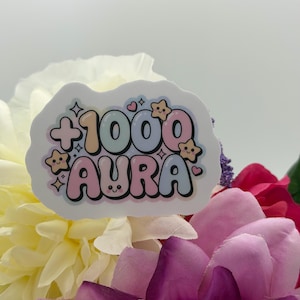 May include: A die-cut sticker featuring the text +1000 AURA in a bubbly, multi-colored font. The design includes small, smiling star icons and hearts in pastel shades of pink, blue, and yellow, with a white border surrounding the entire graphic.