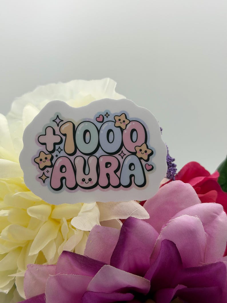 May include: A die-cut sticker featuring the text +1000 AURA in a bubbly, multi-colored font. The design includes small, smiling star icons and hearts in pastel shades of pink, blue, and yellow, with a white border surrounding the entire graphic.