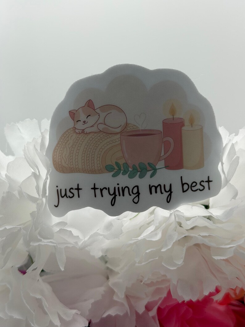 K&ouml;nnte beinhalten: Ein gestanzter Aufkleber mit einer schlafenden Katze auf einer beigen Strickdecke, einer rosa Tasse, zwei brennenden Kerzen und gr&uuml;nen Bl&auml;ttern. Der Text "just trying my best" ist in schwarzer Schreibschrift unten aufgedruckt.