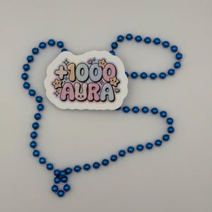 May include: A sticker featuring the text +1000 AURA in pastel bubble letters with small star and heart accents, surrounded by a string of shiny, metallic blue spherical beads arranged in a loose, open loop shape on a plain background.