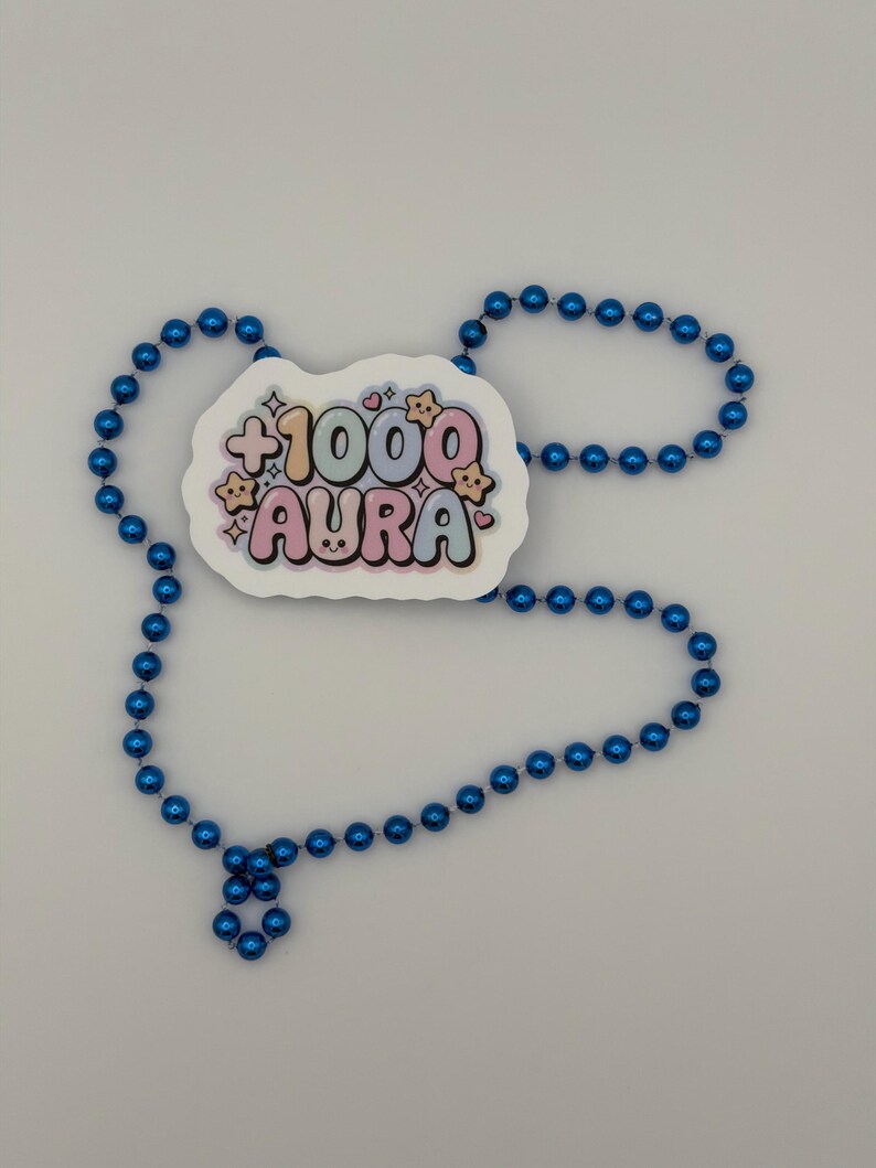 May include: A sticker featuring the text +1000 AURA in pastel bubble letters with small star and heart accents, surrounded by a string of shiny, metallic blue spherical beads arranged in a loose, open loop shape on a plain background.