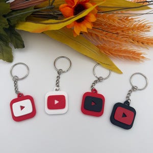May include: Four square keychains featuring a rounded-corner design with a central play button icon. The keychains are rendered in various color combinations including red and white, white and red, red and black, and black and red.