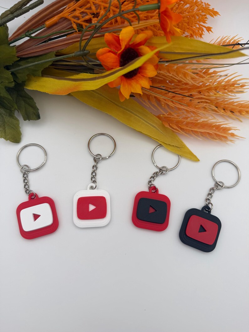 May include: Four square keychains featuring a rounded-corner design with a central play button icon. The keychains are rendered in various color combinations including red and white, white and red, red and black, and black and red.