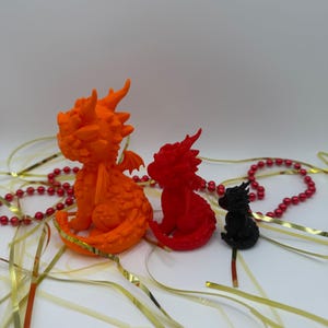 May include: Three dragon figurines in descending sizes, colored orange, red, and black. Each dragon features textured scales, small wings, and horns. The figures are arranged in a row, accompanied by scattered gold ribbons and a string of red beads.