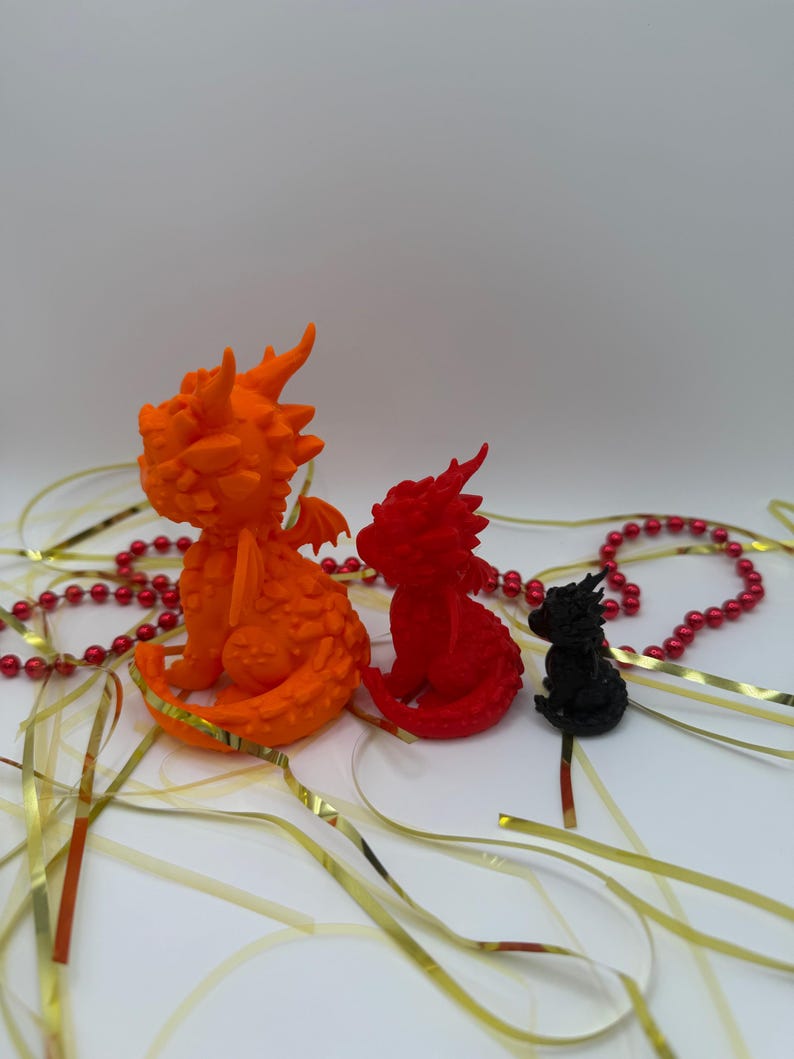 May include: Three dragon figurines in descending sizes, colored orange, red, and black. Each dragon features textured scales, small wings, and horns. The figures are arranged in a row, accompanied by scattered gold ribbons and a string of red beads.