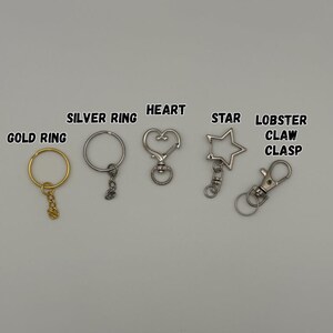 May include: A collection of metal keychain hardware including a gold ring, a silver ring, a heart-shaped clasp, a star-shaped clasp, and a lobster claw clasp. Each item is labeled with its corresponding name in black text above the hardware.