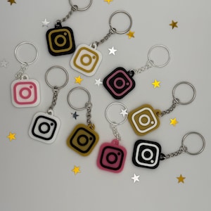 May include: A collection of square keychains featuring the Instagram camera icon in various color combinations, including black, white, pink, and gold. Each keychain is attached to a silver-toned metal split ring by a short chain.