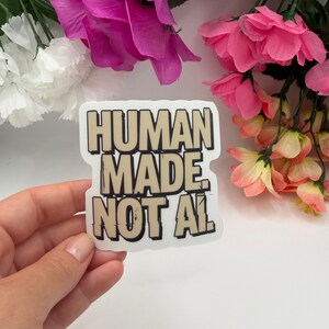 May include: A die-cut sticker featuring the text "HUMAN MADE NOT AI" in bold, blocky, beige letters with a thick black outline. The lettering has a distressed, textured appearance, and the sticker has a clean white border against a plain background.