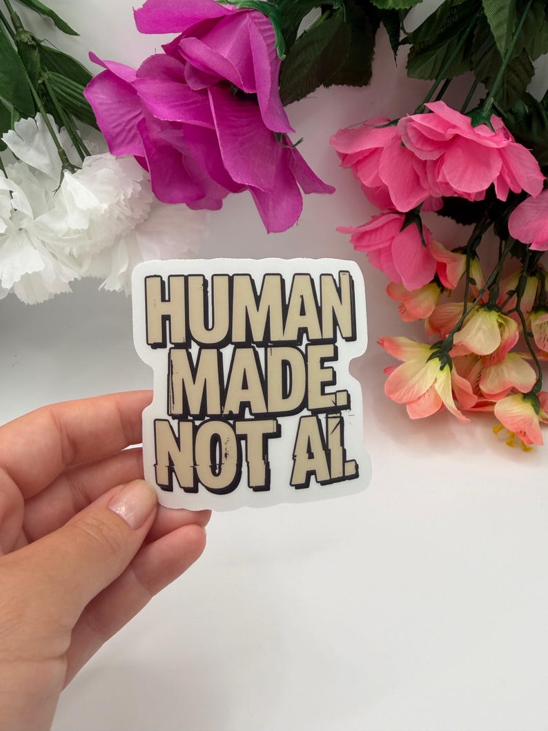 May include: A die-cut sticker featuring the text "HUMAN MADE NOT AI" in bold, blocky, beige letters with a thick black outline. The lettering has a distressed, textured appearance, and the sticker has a clean white border against a plain background.