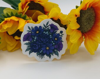 Puede incluir: Una pegatina troquelada que presenta una ilustración detallada de un ramo de flores de color azul oscuro y morado con hojas verdes. La pegatina está colocada frente a dos grandes girasoles de color amarillo brillante con centros marrones.