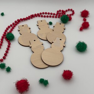 May include: Five flat, light-colored wooden snowman cutouts, each featuring a top hat and a small hole for hanging. The ornaments are arranged together, surrounded by a string of red beads and several small red and green fuzzy pom-poms.