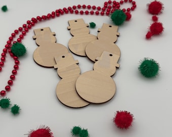 May include: Five flat, light-colored wooden snowman cutouts, each featuring a top hat and a small hole for hanging. The ornaments are arranged together, surrounded by a string of red beads and several small red and green fuzzy pom-poms.