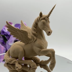 Puede incluir: Una figura de unicornio monocromática de color tostado claro con un cuerno en espiral, alas emplumadas y patrones florales en relieve en el cuerpo. La criatura está sentada con una pata delantera levantada, mostrando texturas detalladas en la crin y la cola.
