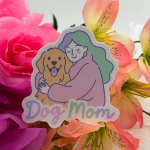 Op de afbeelding: Een gestanste sticker met een lijntekening van een persoon met golvend blauwgroen haar in een roze trui, die een goudkleurige hond knuffelt. De tekst "Dog Mom" staat onderaan in een zacht, afgerond paars lettertype gedrukt.