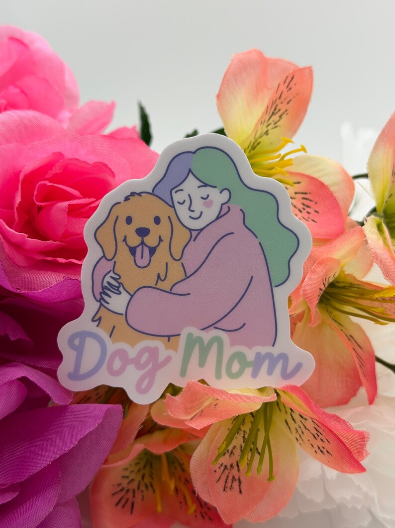 Op de afbeelding: Een gestanste sticker met een lijntekening van een persoon met golvend blauwgroen haar in een roze trui, die een goudkleurige hond knuffelt. De tekst "Dog Mom" staat onderaan in een zacht, afgerond paars lettertype gedrukt.