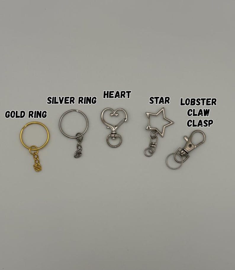 May include: A collection of metal keychain hardware including a gold ring, a silver ring, a heart-shaped clasp, a star-shaped clasp, and a lobster claw clasp. Each item is labeled with its corresponding name in black text above the hardware.