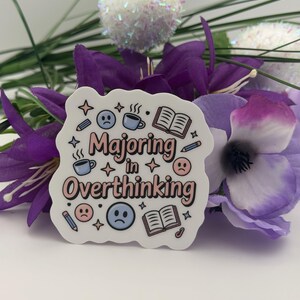 May include: A die-cut sticker with a white background featuring the text "Majoring in Overthinking" in pink and black. The design includes illustrations of blue coffee mugs, open books, pencils, four-pointed stars, and sad blue and pink faces.