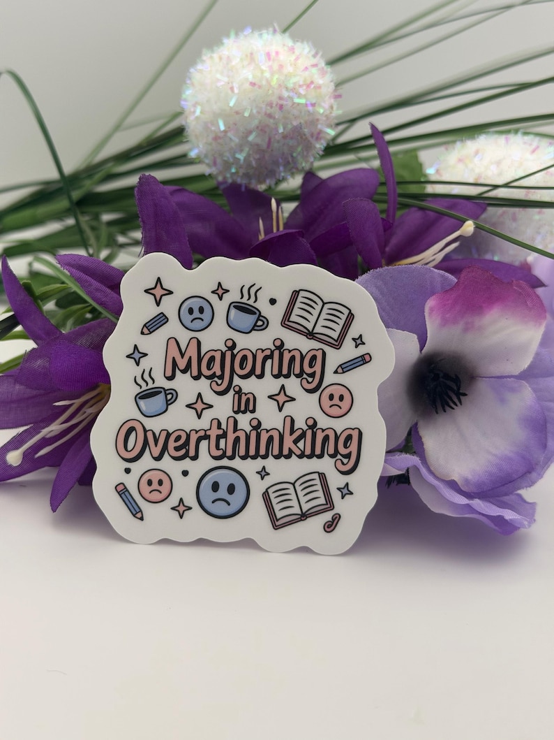 May include: A die-cut sticker with a white background featuring the text "Majoring in Overthinking" in pink and black. The design includes illustrations of blue coffee mugs, open books, pencils, four-pointed stars, and sad blue and pink faces.