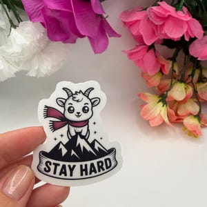 May include: A white die-cut sticker featuring a cartoon goat wearing a maroon scarf, standing atop a black mountain peak. The design includes small star accents and a banner at the bottom with the black text STAY HARD printed in a bold font.