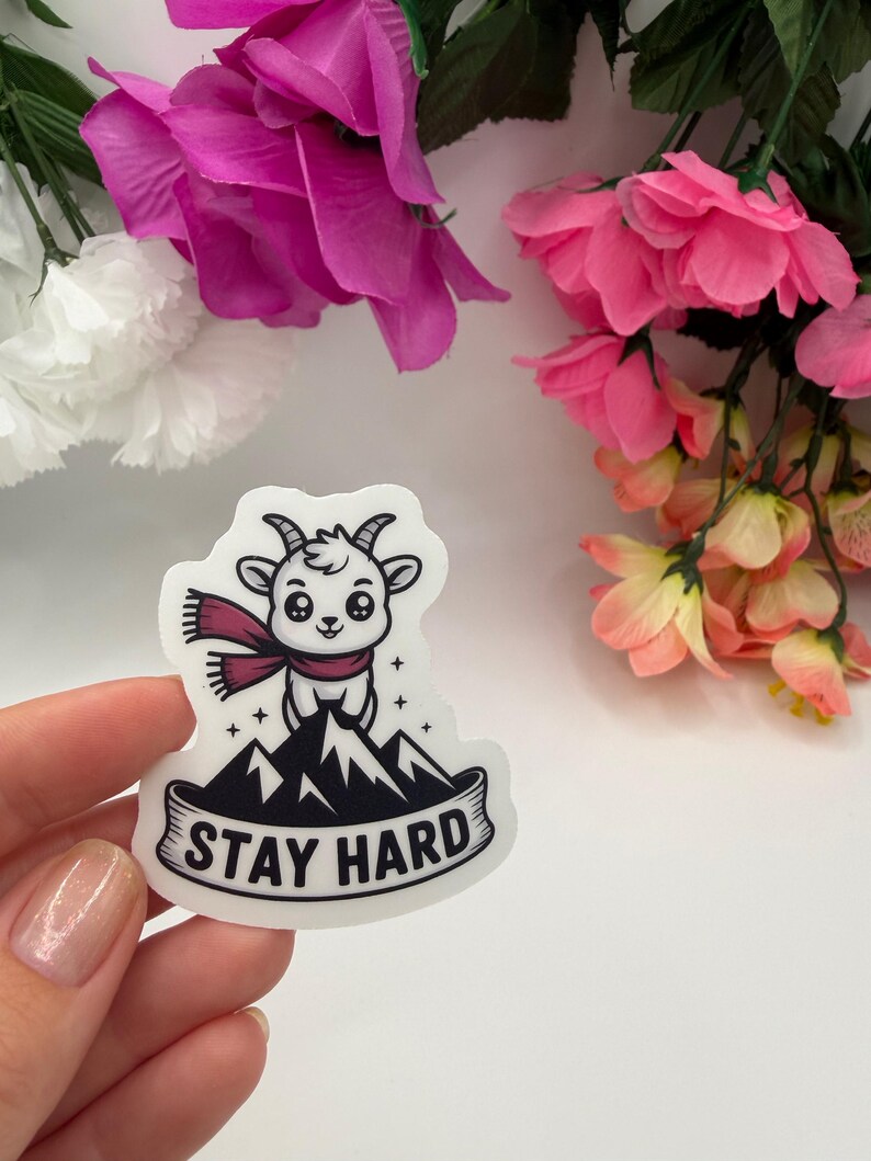 May include: A white die-cut sticker featuring a cartoon goat wearing a maroon scarf, standing atop a black mountain peak. The design includes small star accents and a banner at the bottom with the black text STAY HARD printed in a bold font.