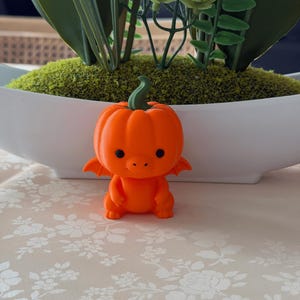 May include: A small, bright orange figurine shaped like a pumpkin with a green stem on top. The figure has a rounded body, two small wings on the sides, and a smooth surface with two black circular eyes and a small snout on the front face.
