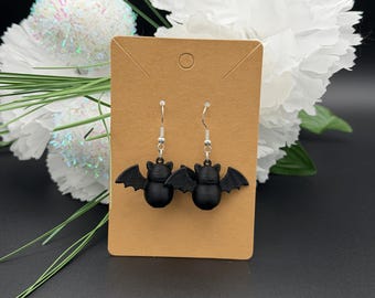 May include: A pair of matte black bat-shaped earrings featuring a rounded body and outstretched wings. The earrings are attached to silver-toned metal hooks and are displayed on a rectangular tan cardboard jewelry card with a hole at the top.